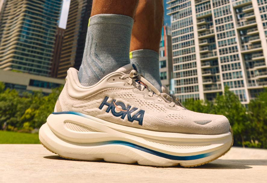 Gros plan d’une personne portant des chaussures de course HOKA avec une semelle épaisse et rembourrée, debout à l’extérieur avec des bâtiments urbains en arrière-plan.