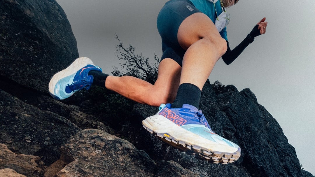 Coureur portant des chaussures bleues HOKA de course sur sentier en train de naviguer sur un sentier rocheux abrupt en plein air.