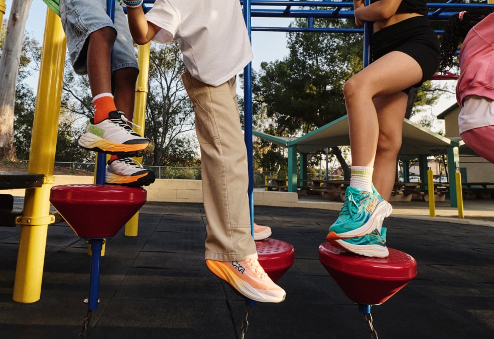 Les enfants jouent sur une structure d'escalade dans une aire de jeux, mettant en valeur des chaussures de course HOKA colorées et des vêtements de sport décontractés.
