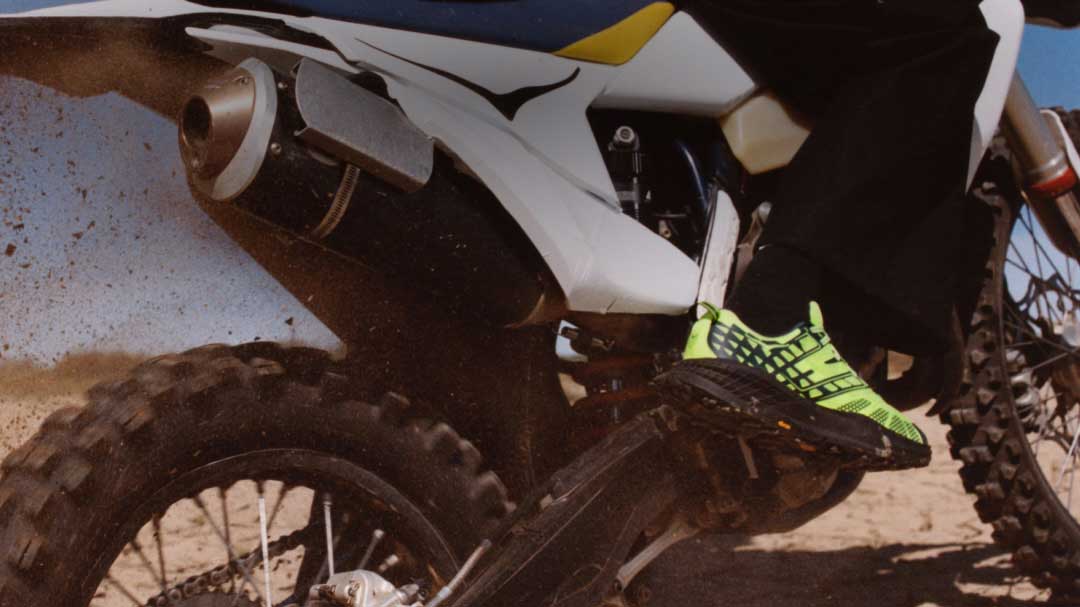 Gros plan d’une moto de crosse soulevant la terre sur une piste extérieure, avec un cycliste portant des chaussures de course vert fluo.