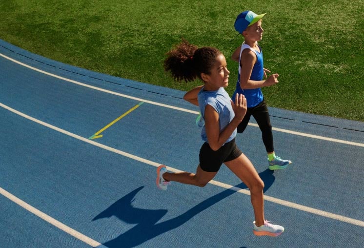 Two kids running on an outdoor track, wearing HOKA shoes.
