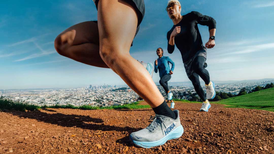 Un gros plan des jambes d’un coureur en mouvement sur un sentier de terre, suivi par deux autres coureurs sur fond de skyline urbain.
