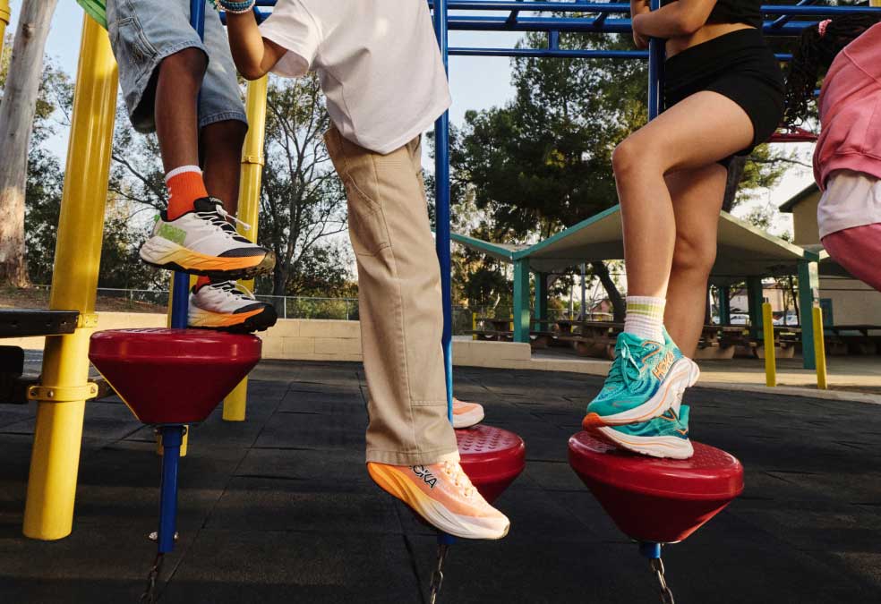 Les enfants jouent sur une structure d'escalade dans une aire de jeux, mettant en valeur des chaussures de course HOKA colorées et des vêtements de sport décontractés.