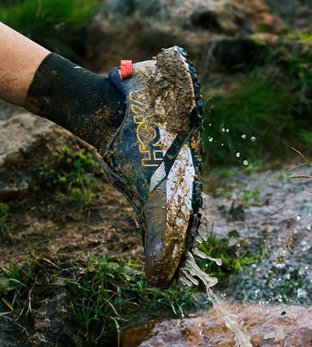 Une personne courant dans un ruisseau, portant des chaussures HOKA mouillées et boueuses.