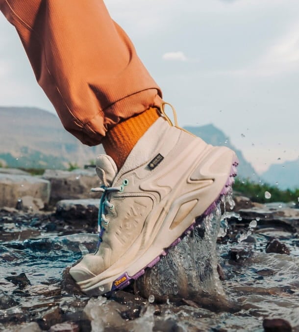 Personne qui entre dans l’eau rocailleuse en chaussures HOKA.