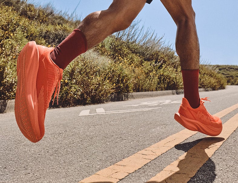 Coureur portant des chaussures de course orange vif HOKA sur une route ensoleillée lors d’une course en plein air.