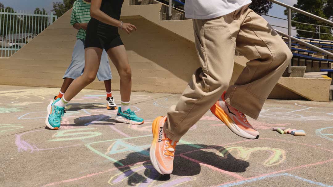 Gros plan d'enfants jouant à la marelle sur un plancher de béton recouvert de motifs colorés à la craie, portant des espadrilles HOKA orange et turquoise.
