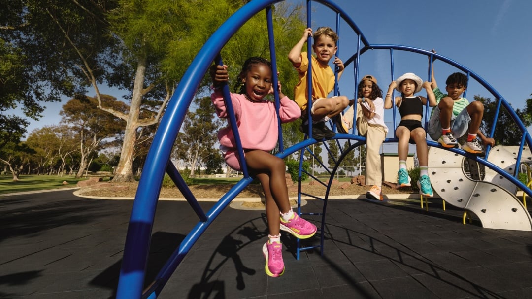 Cinq enfants souriants, chaussés de chaussures HOKA colorées, grimpent sur une structure de jeu bleue en forme d'arche, dans un parc ensoleillé.