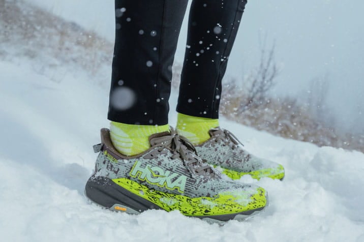 Personne debout sur une colline enneigée, portant des chaussures et de l'équipement HOKA